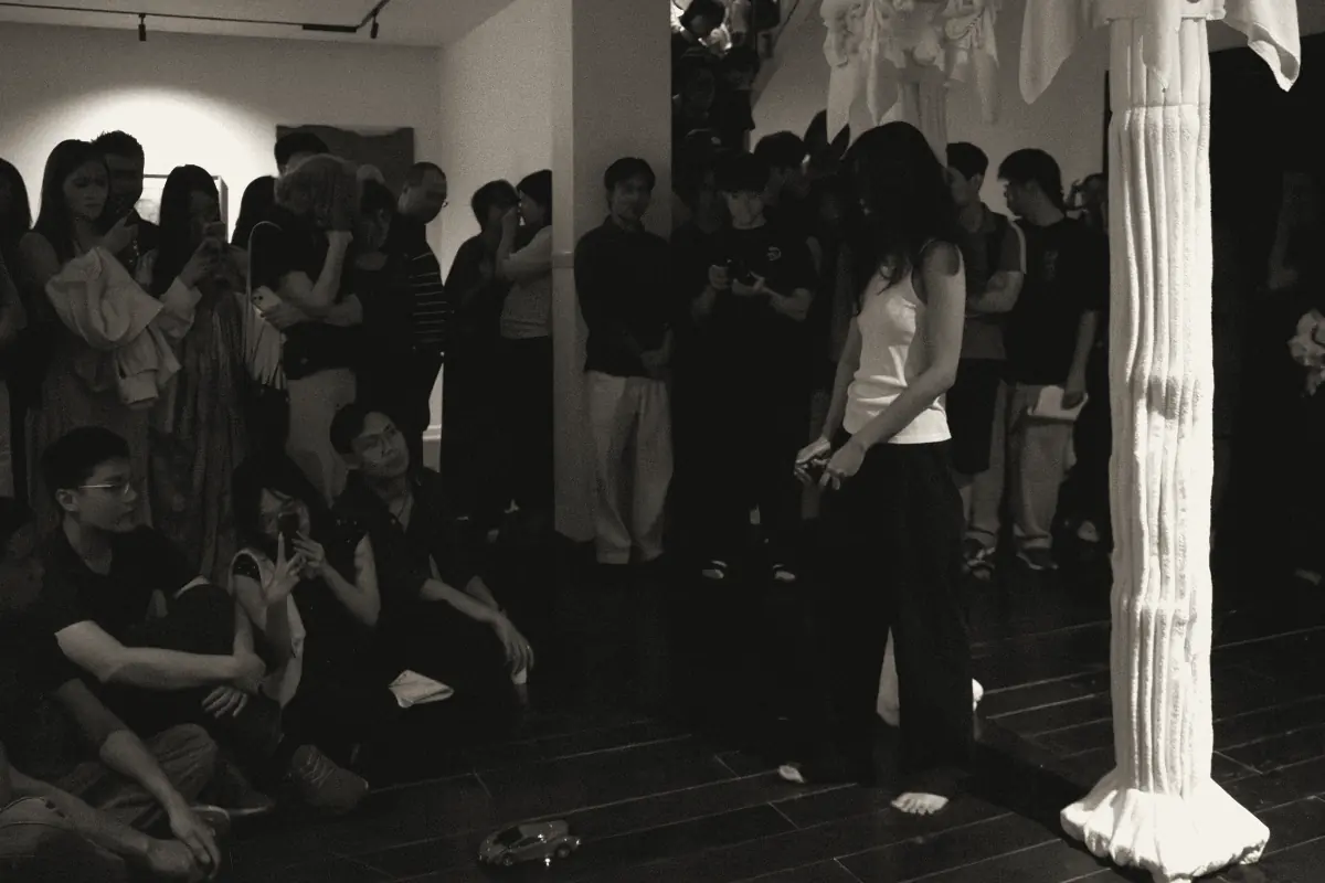A performance in a gallery, the audience seated and standing around the performer, a toy car on the floor